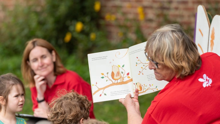 Volunteer reading a story from a book at a summer event with children sat on the grass listening